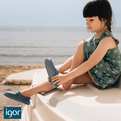 Criança com sapatos tipo crocs azuis e vestido verde com padrão de abacaxis sentada na praia
