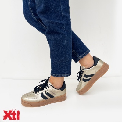 Ténis dourados e pretos da marca Xti usados por uma pessoa com jeans azul
