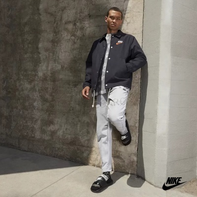 Homem com casaco azul escuro e calças cinza vestindo chinelos pretos Nike encostado em parede cinzenta.