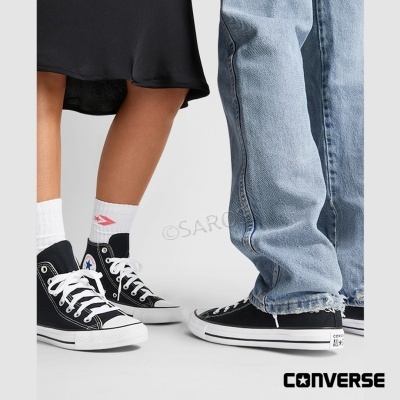 Ténis Converse All Star pretos com atacadores brancos, usados com meias e calças de ganga, fundo branco logo CONVERSE.