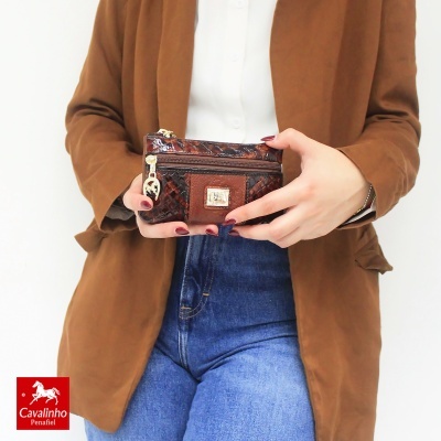 Carteira pequena de couro marrom com padrão entrelaçado e fecho dourado das mãos de uma mulher com casaco castanho e jeans azuis