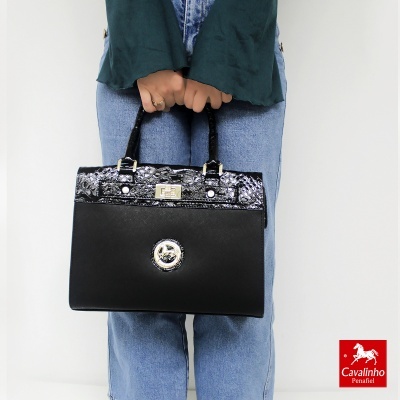 Mala preta com padrão de pele de cobra e alças pretas, modelo segura com as duas mãos, jeans azuis e camisa verde