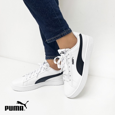 Ténis Puma brancos com detalhes em preto usados com calça jeans azul escura em fundo branco