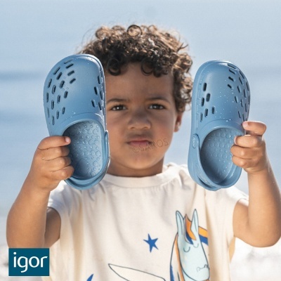 Criança com sapatos tipo crocs azuis na mão vestindo t-shirt branca com estampa de cavalo.
