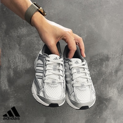 Par de sapatilhas Adidas brancas e cinzentas seguradas por uma mão em fundo cinzento.