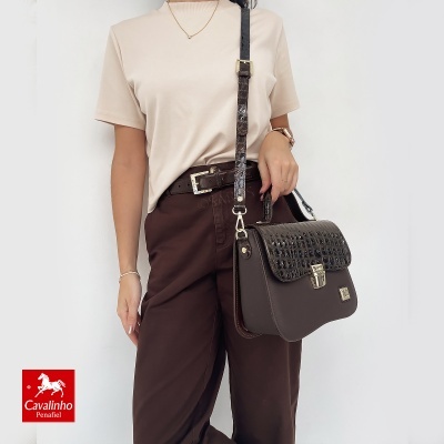 Mulher com mala de ombro castanha escura com textura crocodilo e fivela dourada, vestindo calças castanhas e t-shirt bege