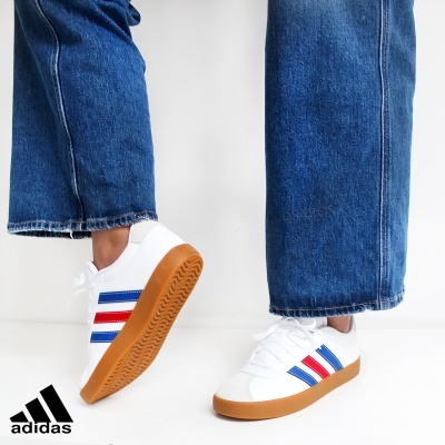 Ténis Adidas brancos com riscas azuis e vermelhas e sola castanha em piso branco