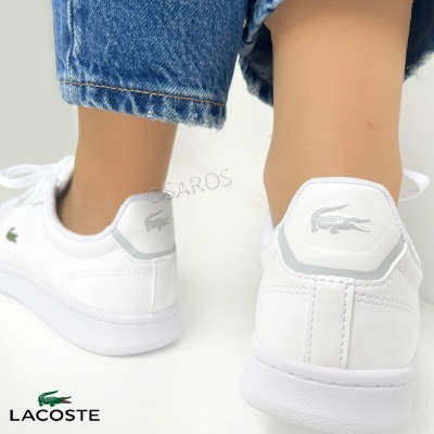 Sapatilhas brancas Lacoste com logo crocodilo cinza na parte traseira, usadas com calças de ganga azul.