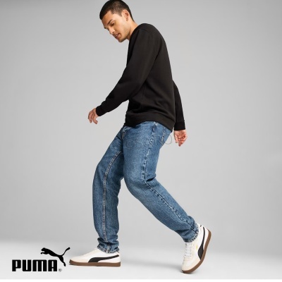 Homem com sweat preto, jeans azul e ténis brancos Puma em fundo cinza.
