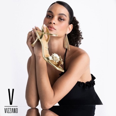 Sandália de salto alto dourada com flor frontal segurada por mulher com vestido preto e brinco dourado