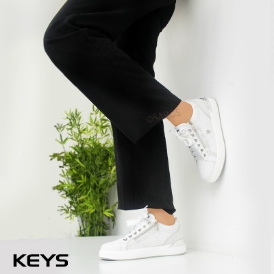 Ténis brancos com padrão crocodilo e tachas, calças pretas e planta verde ao fundo