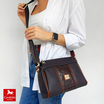 Mala de ombro castanha e castanho escuro com detalhes dourados, usada por mulher com camisa branca e jeans azuis.