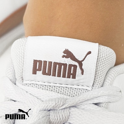 Etiqueta branca com logótipo PUMA castanho num calçado desportivo branco.