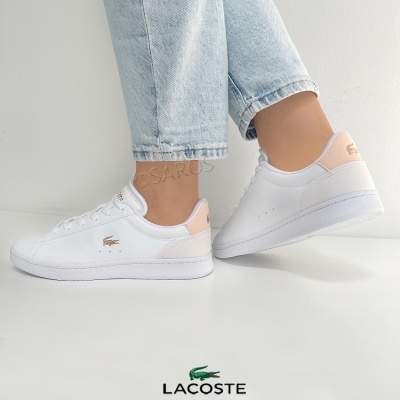 Ténis brancos com detalhes rosa claro e logo Lacoste em fundo branco