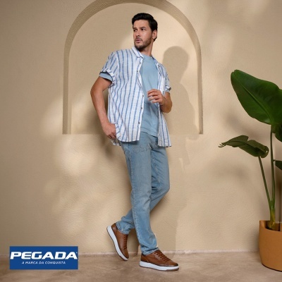 Homem com calças de ganga azuis claras, t-shirt azul claro, camisa às riscas branca e azul, sapatos castanhos, planta verde em vaso castanho com fundo bege