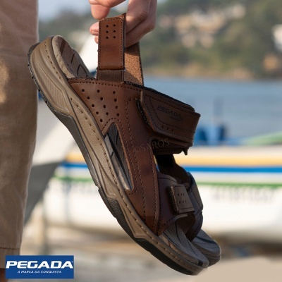 Sandália castanha masculina com tiras de velcro e sola robusta, numa mão, com fundo de barco e água