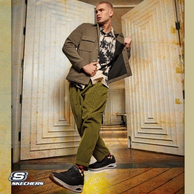 Homem com ténis Sketchers pretos, calças verdes, camisa estampada e casaco castanho interior