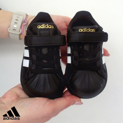 Ténis infantis Adidas pretos com faixas brancas e logo dourado, segurados por mãos femininas