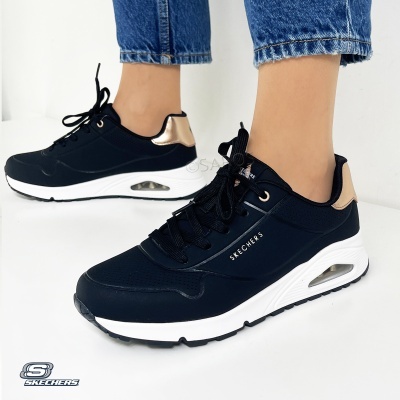 Ténis pretos Skechers com detalhes dourados e sola branca, usados com calças de ganga azuis