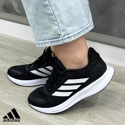 Ténis Adidas pretos com riscas brancas e sola branca, usados com calças de ganga claras em chão de madeira clara
