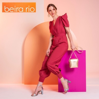 Mulher com macacão vermelho, sapatos prateados e mala dourada em fundo geométrico rosa e laranja