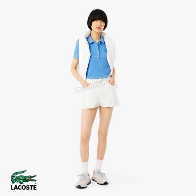 Mulher com polo azul, calções brancos e sapatilhas em fundo branco com logo Lacoste