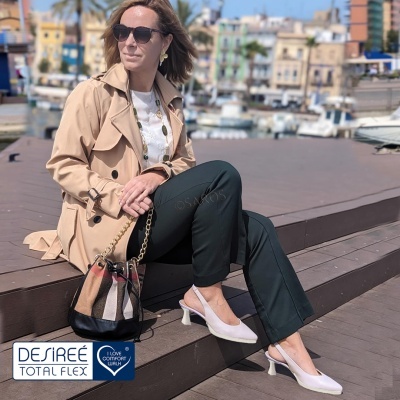 Mulher com casaco bege e sapatos brancos sentada num cais com barcos ao fundo