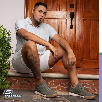 Homem com sapatilhas verdes Skechers, camiseta e calções brancos, sentado à porta de madeira