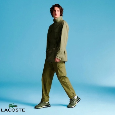 Homem vestindo casaco e calças verdes com calçado desportivo, fundo azul, logótipo Lacoste.