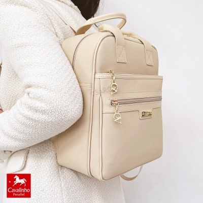 Mochila bege de material sintético com detalhes dourados e placa metálica Cavalinho, usada por pessoa com casaco creme.