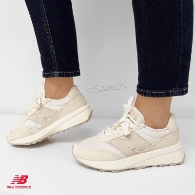 Ténis New Balance bege com sola branca e atacadores, usados por pessoa com calças pretas.
