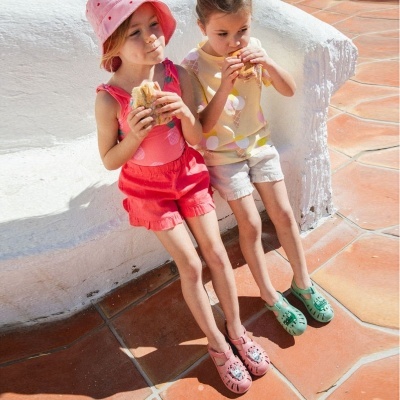 Duas crianças sentadas num banco branco, usam roupa colorida e sapatos rosa e verde, comem sandes num piso de ladrilho terracota.