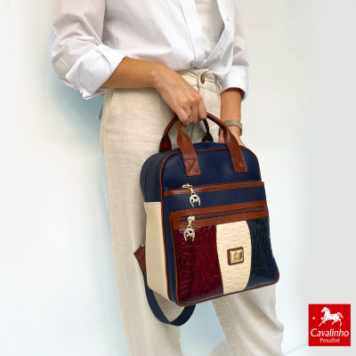 Mochila Cavalinho Penafiel azul, bege, vinho e castanha com detalhes em pele e fechos dourados, segurada por pessoa com camisa branca e calças bege.