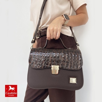 Mala castanha escura com tampa texturizada tipo crocodilo e fecho dourado, usada por uma mulher.