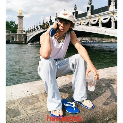 Pessoa com chinelos Havaianas azuis e roupa casual junto a um rio, com ponte e estátua ao fundo