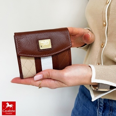 Carteira Cavalinho castanha em couro com detalhe em tecido bege, com placa dourada, segurada por mão humana.