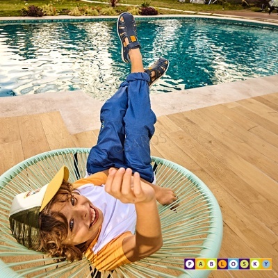 Menino de calças azuis e t-shirt branca com mangas amarelo-alaranjadas sentado numa cadeira verde claro junto a uma piscina.