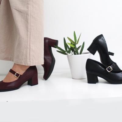 Dois pares de sapatos Mary Jane em bordô e preto com saltos médios e fivelas, junto a uma planta num vaso branco.