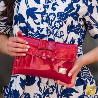 Carteira vermelha brilhante com laço, segurada em mãos sobre camisa branca com padrão azul