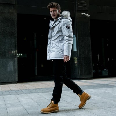 Homem com casaco branco de capuz peludo, calças pretas e botas castanhas em ambiente urbano.