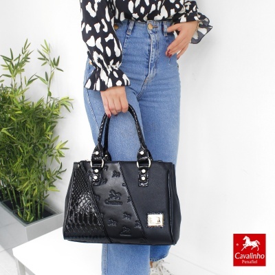 Mala preta de senhora com detalhes em relevo e padrão texturizado, segurada por mulher com camisa preta e jeans azuis