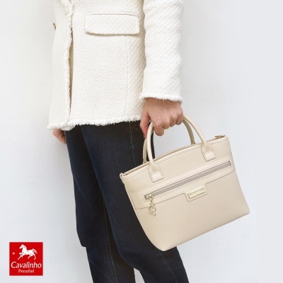 Bolsa de mão bege com fecho dourado na mão de pessoa com casaco branco e calças pretas