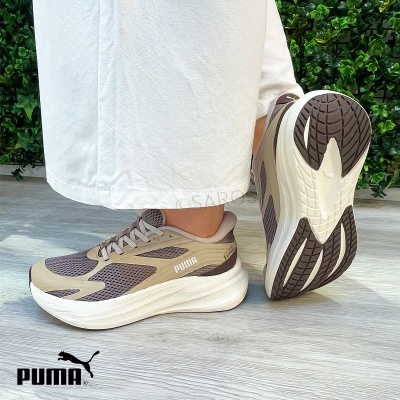 Sapatos desportivos PUMA bege e castanho com sola branca usados por uma pessoa em chão cinza claro e fundo de plantas verdes