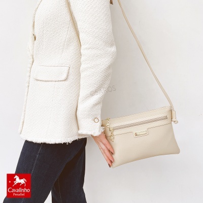 Mala tiracolo bege claro com detalhes metálicos dourados junto a mulher com casaco branco.