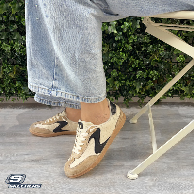 Ténis Skechers bege, branco e preto com sola castanha, texturas matelassê e camurça, junto a cadeira creme e plantas verdes