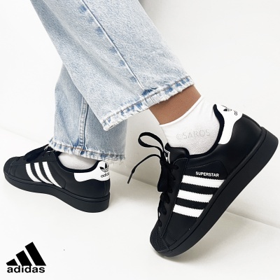 Ténis Adidas Superstar pretos com riscas brancas usados com meias brancas e calças de ganga azul clara