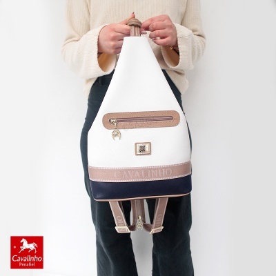 Mochila triangular branca, bege e azul escuro com alças e bolso frontal, segurada por uma pessoa.