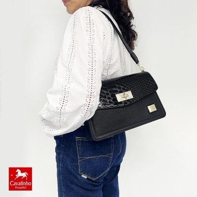 Mala de senhora preta com padrão de pele de crocodilo e fecho dourado, alça preta, usada por mulher com camisa branca bordada e jeans azul.