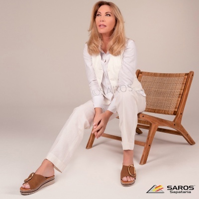 Mulher sentada numa cadeira de madeira com assento entrelaçado, veste roupa branca e sandálias castanhas com fivela dourada