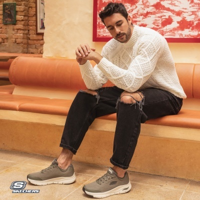 Homem sentado com camisola branca, jeans pretos rasgados e sapatilhas cinzentas Skechers